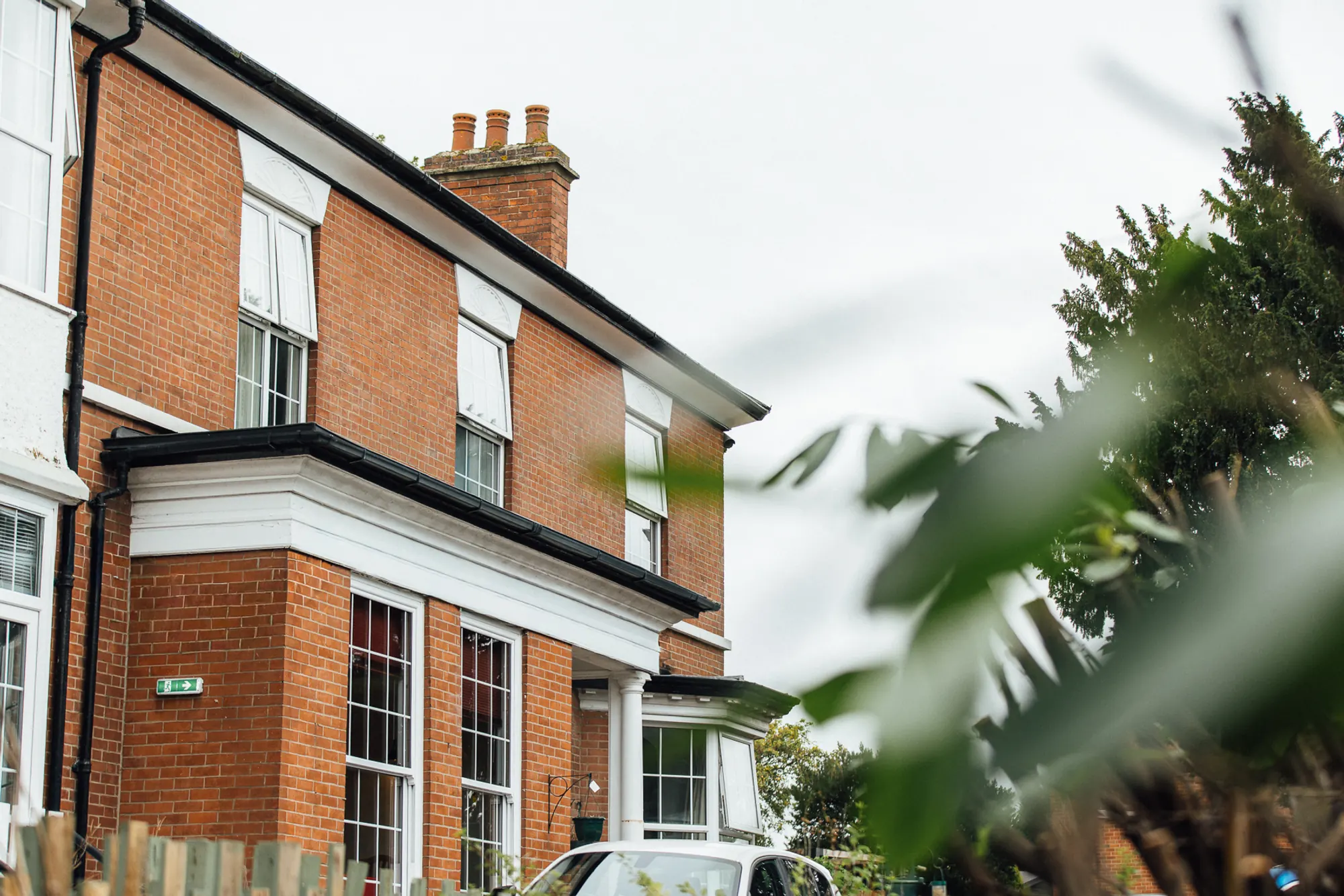 The red-brick front of Eastwood House, with white sash windows and a white porch, seen past garden leaves