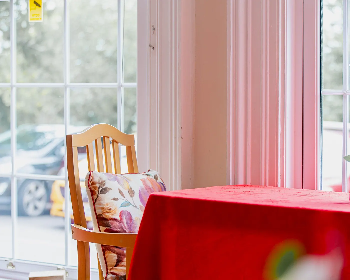 Dining table with a red cloth and a wooden chair with a floral cushion by the window